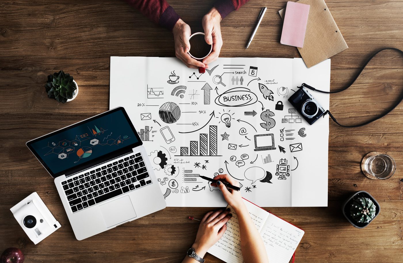 Hands sketching a business plan beside a laptop showing analytics charts